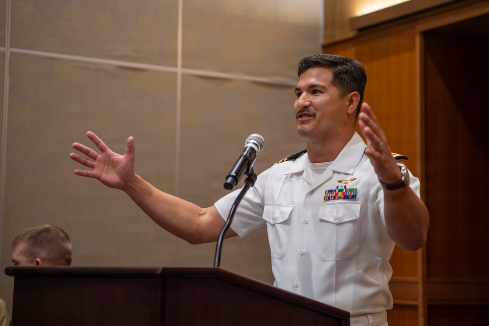 A U.S. Navy officer in white service dress uniform is speaking at a podium with a microphone, gesturing expressively with both hands raised. He is wearing multiple service ribbons, a naval aviator or officer badge, and rank insignia on his shoulders. The setting appears to be an indoor formal or ceremonial event.