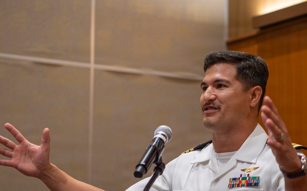 A U.S. Navy officer in white service dress uniform is speaking at a podium with a microphone, gesturing expressively with both hands raised. He is wearing multiple service ribbons, a naval aviator or officer badge, and rank insignia on his shoulders. The setting appears to be an indoor formal or ceremonial event.