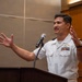 A U.S. Navy officer in white service dress uniform is speaking at a podium with a microphone, gesturing expressively with both hands raised. He is wearing multiple service ribbons, a naval aviator or officer badge, and rank insignia on his shoulders. The setting appears to be an indoor formal or ceremonial event.