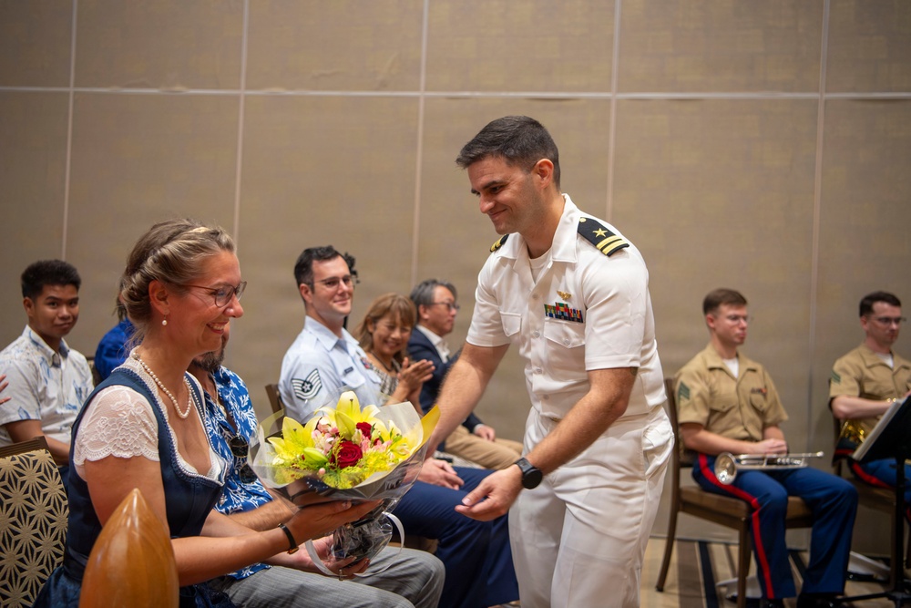 A naval officer in white dress uniform is presenting a bouquet of flowers to a seated woman wearing glasses and a pearl necklace at what appears to be a formal military ceremony or event. The audience behind them includes personnel in various military uniforms including Air Force and Marine Corps, as well as civilians. The atmosphere appears celebratory and warm.