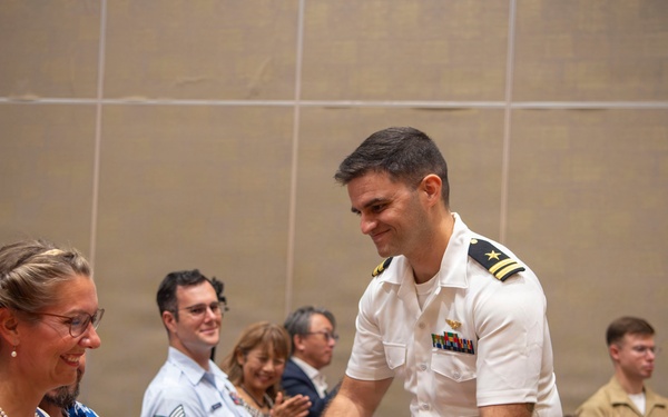 A naval officer in white dress uniform is presenting a bouquet of flowers to a seated woman wearing glasses and a pearl necklace at what appears to be a formal military ceremony or event. The audience behind them includes personnel in various military uniforms including Air Force and Marine Corps, as well as civilians. The atmosphere appears celebratory and warm.