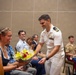 A naval officer in white dress uniform is presenting a bouquet of flowers to a seated woman wearing glasses and a pearl necklace at what appears to be a formal military ceremony or event. The audience behind them includes personnel in various military uniforms including Air Force and Marine Corps, as well as civilians. The atmosphere appears celebratory and warm.