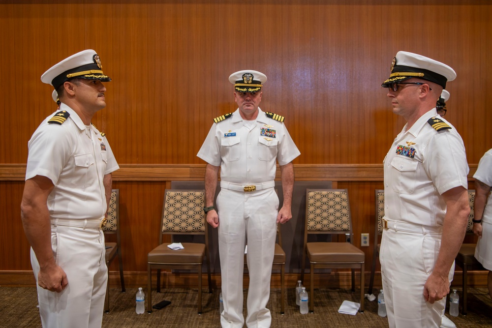 Three U.S. Navy officers in white dress uniforms are standing in what appears to be a formal indoor setting, likely during a military ceremony or change of command event. Two officers face each other in the foreground while a third stands between them in the background. The setting includes chairs arranged along a wood-paneled wall, suggesting a conference room or ceremonial hall.