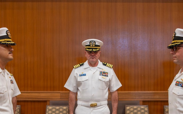 Three U.S. Navy officers in white dress uniforms are standing in what appears to be a formal indoor setting, likely during a military ceremony or change of command event. Two officers face each other in the foreground while a third stands between them in the background. The setting includes chairs arranged along a wood-paneled wall, suggesting a conference room or ceremonial hall.