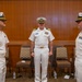 Three U.S. Navy officers in white dress uniforms are standing in what appears to be a formal indoor setting, likely during a military ceremony or change of command event. Two officers face each other in the foreground while a third stands between them in the background. The setting includes chairs arranged along a wood-paneled wall, suggesting a conference room or ceremonial hall.