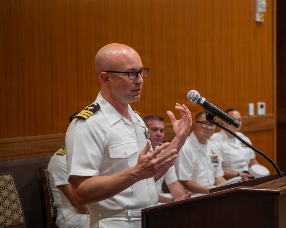 A bald male naval officer wearing a white dress uniform with gold rank insignia on the shoulder boards is speaking at a wooden podium equipped with a microphone. He is gesturing with both hands while addressing an audience. Several other uniformed naval personnel are seated in the background.