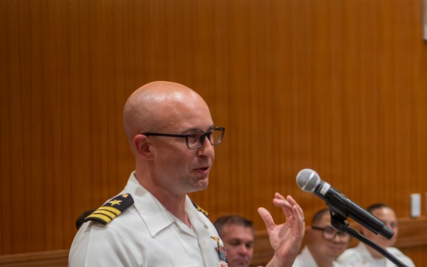 A bald male naval officer wearing a white dress uniform with gold rank insignia on the shoulder boards is speaking at a wooden podium equipped with a microphone. He is gesturing with both hands while addressing an audience. Several other uniformed naval personnel are seated in the background.