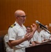 A bald male naval officer wearing a white dress uniform with gold rank insignia on the shoulder boards is speaking at a wooden podium equipped with a microphone. He is gesturing with both hands while addressing an audience. Several other uniformed naval personnel are seated in the background.