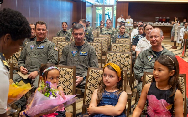 A formal military ceremony is taking place in a decorated hall with a red carpet aisle. Three young girls dressed in formal attire are seated in the front row, with one receiving a bouquet of flowers from a person in a white naval uniform. Military personnel in various uniforms, including olive flight suits and white dress uniforms, are seated in the audience.