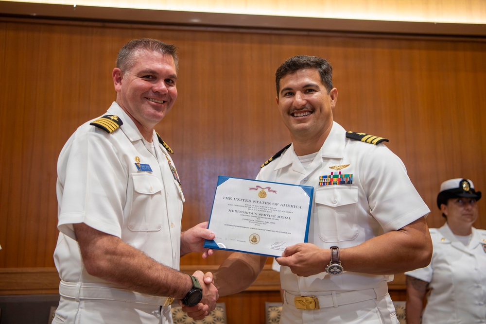 Two U.S. Navy officers in white service dress uniforms are shaking hands while jointly holding a Meritorious Service Medal certificate issued by The United States of America. The ceremony appears to be taking place in a formal indoor setting with wood-paneled walls. A third uniformed Navy personnel member is visible in the background.