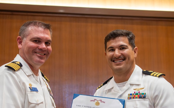 Two U.S. Navy officers in white service dress uniforms are shaking hands while jointly holding a Meritorious Service Medal certificate issued by The United States of America. The ceremony appears to be taking place in a formal indoor setting with wood-paneled walls. A third uniformed Navy personnel member is visible in the background.