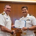 Two U.S. Navy officers in white service dress uniforms are shaking hands while jointly holding a Meritorious Service Medal certificate issued by The United States of America. The ceremony appears to be taking place in a formal indoor setting with wood-paneled walls. A third uniformed Navy personnel member is visible in the background.