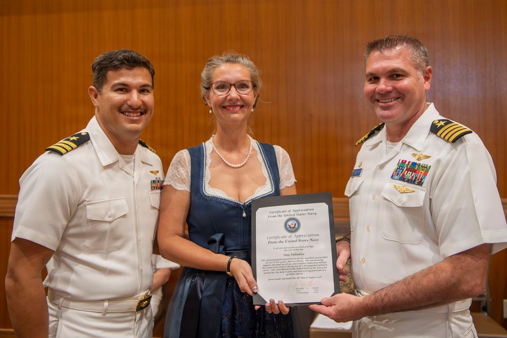 Three individuals pose together holding a Certificate of Appreciation from the United States Navy. Two men in white U.S. Navy dress uniforms flank a woman in a blue dress who is holding the framed certificate. The setting appears to be an indoor formal ceremony or official event.
