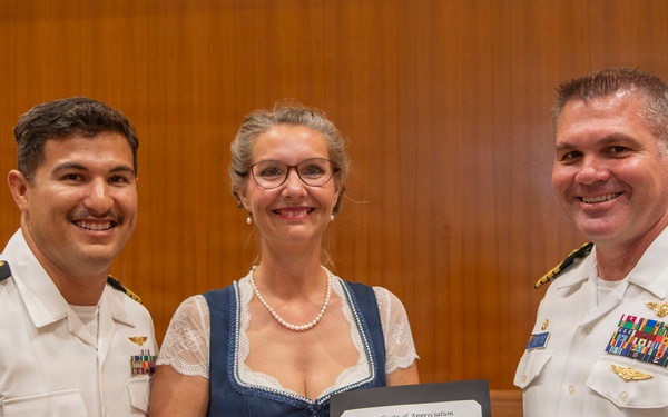 Three individuals pose together holding a Certificate of Appreciation from the United States Navy. Two men in white U.S. Navy dress uniforms flank a woman in a blue dress who is holding the framed certificate. The setting appears to be an indoor formal ceremony or official event.