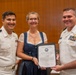 Three individuals pose together holding a Certificate of Appreciation from the United States Navy. Two men in white U.S. Navy dress uniforms flank a woman in a blue dress who is holding the framed certificate. The setting appears to be an indoor formal ceremony or official event.