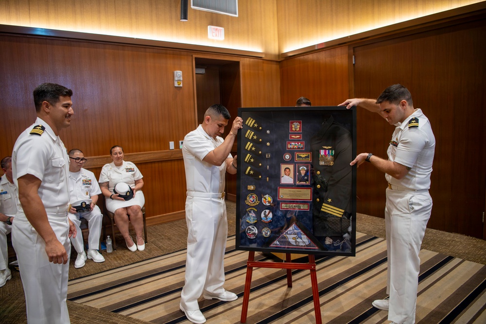 A U.S. Navy retirement or recognition ceremony is taking place in a wood-paneled room. Three naval officers in white dress uniforms are presenting a shadow box display case containing medals, patches, rank insignia, photographs, and a folded flag. Seated personnel in the background observe the presentation.