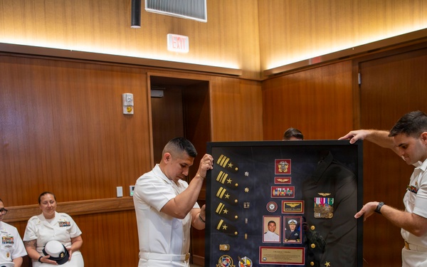 A U.S. Navy retirement or recognition ceremony is taking place in a wood-paneled room. Three naval officers in white dress uniforms are presenting a shadow box display case containing medals, patches, rank insignia, photographs, and a folded flag. Seated personnel in the background observe the presentation.