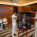 A U.S. Navy retirement or recognition ceremony is taking place in a wood-paneled room. Three naval officers in white dress uniforms are presenting a shadow box display case containing medals, patches, rank insignia, photographs, and a folded flag. Seated personnel in the background observe the presentation.