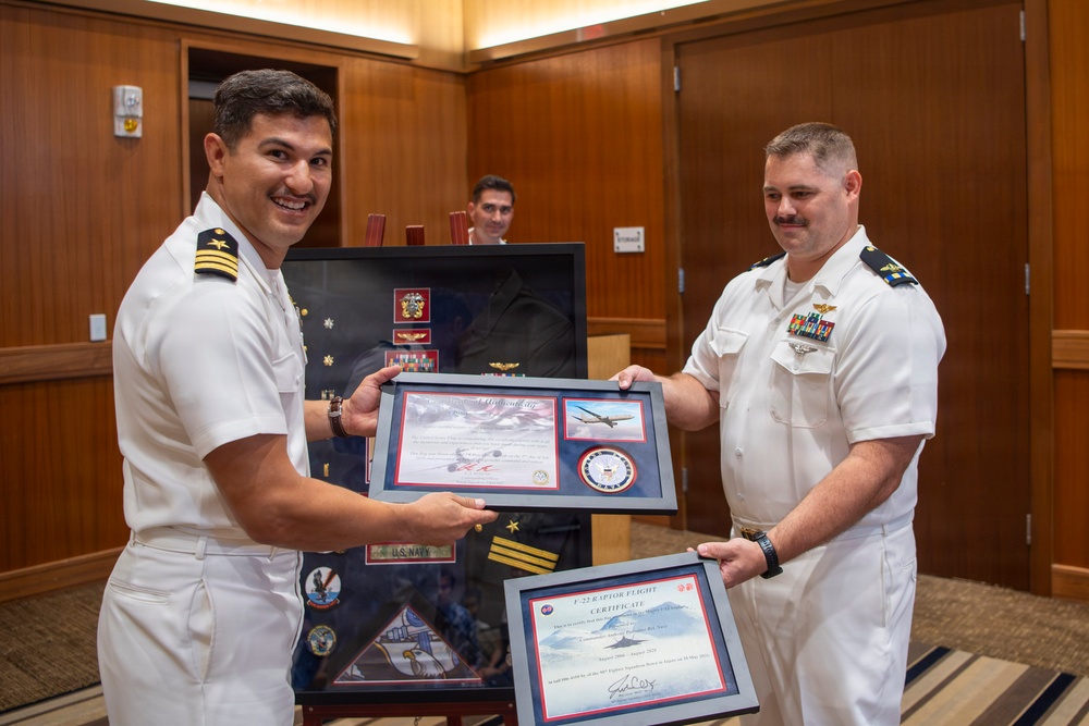 Two U.S. Navy officers in white service uniforms are jointly presenting or receiving framed certificates and awards during what appears to be a formal military ceremony. The officer on the left is smiling broadly while both hold a framed display containing what appears to be an F-22 Raptor Flight Certificate along with other military memorabilia. A shadow box containing U.S. Navy insignia, patches, medals, and a folded flag is visible in the background.