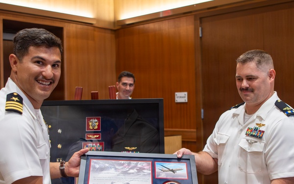 Two U.S. Navy officers in white service uniforms are jointly presenting or receiving framed certificates and awards during what appears to be a formal military ceremony. The officer on the left is smiling broadly while both hold a framed display containing what appears to be an F-22 Raptor Flight Certificate along with other military memorabilia. A shadow box containing U.S. Navy insignia, patches, medals, and a folded flag is visible in the background.