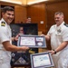 Two U.S. Navy officers in white service uniforms are jointly presenting or receiving framed certificates and awards during what appears to be a formal military ceremony. The officer on the left is smiling broadly while both hold a framed display containing what appears to be an F-22 Raptor Flight Certificate along with other military memorabilia. A shadow box containing U.S. Navy insignia, patches, medals, and a folded flag is visible in the background.