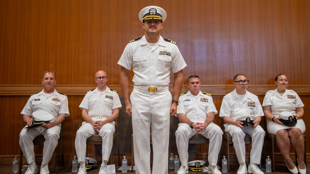 A group of six U.S. Navy personnel dressed in white service uniforms are gathered in what appears to be a formal ceremony or official event. One officer stands at attention in the center foreground, wearing a combination cover (white cap) with gold insignia, while five others are seated behind him. The setting appears to be an indoor venue with wood-paneled walls.