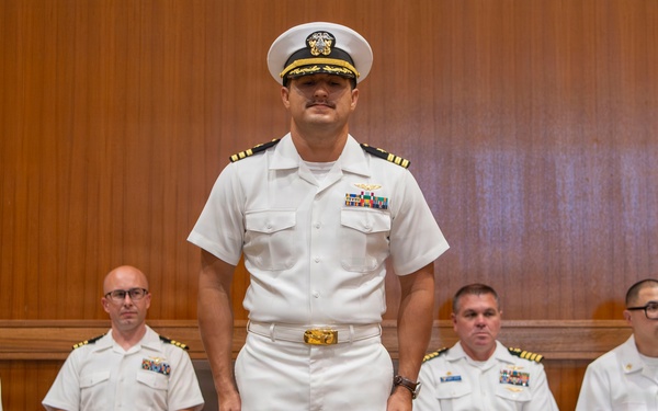 A group of six U.S. Navy personnel dressed in white service uniforms are gathered in what appears to be a formal ceremony or official event. One officer stands at attention in the center foreground, wearing a combination cover (white cap) with gold insignia, while five others are seated behind him. The setting appears to be an indoor venue with wood-paneled walls.