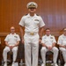 A group of six U.S. Navy personnel dressed in white service uniforms are gathered in what appears to be a formal ceremony or official event. One officer stands at attention in the center foreground, wearing a combination cover (white cap) with gold insignia, while five others are seated behind him. The setting appears to be an indoor venue with wood-paneled walls.