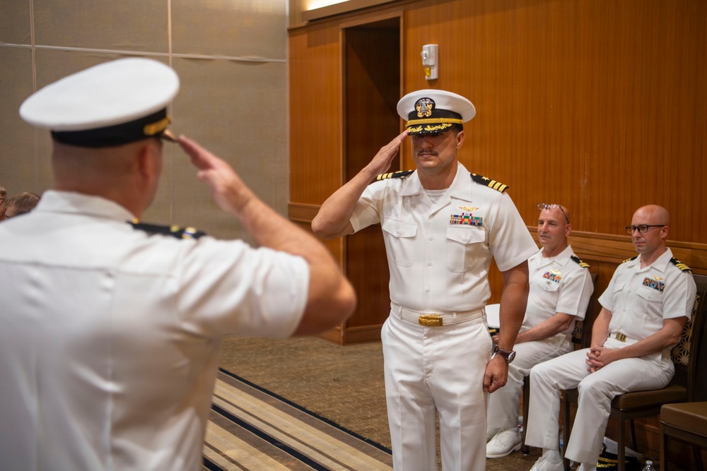 Two U.S. Navy officers in white service dress uniforms are exchanging salutes during what appears to be a formal military ceremony, likely a change of command or promotion event. The central officer facing the camera displays captain's rank insignia (four gold stripes) and multiple service ribbons. Two additional naval officers are seated in the background observing the ceremony.