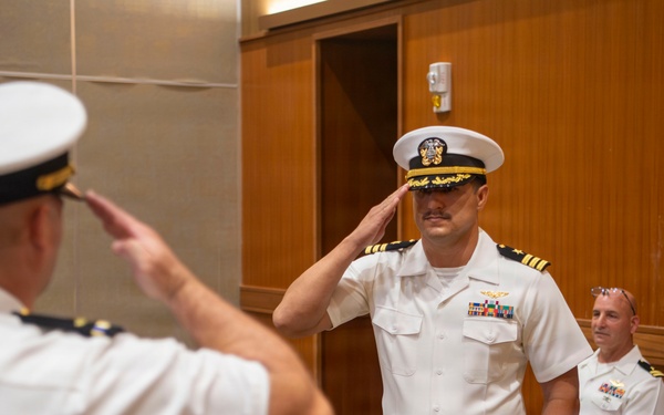 Two U.S. Navy officers in white service dress uniforms are exchanging salutes during what appears to be a formal military ceremony, likely a change of command or promotion event. The central officer facing the camera displays captain's rank insignia (four gold stripes) and multiple service ribbons. Two additional naval officers are seated in the background observing the ceremony.