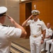 Two U.S. Navy officers in white service dress uniforms are exchanging salutes during what appears to be a formal military ceremony, likely a change of command or promotion event. The central officer facing the camera displays captain's rank insignia (four gold stripes) and multiple service ribbons. Two additional naval officers are seated in the background observing the ceremony.