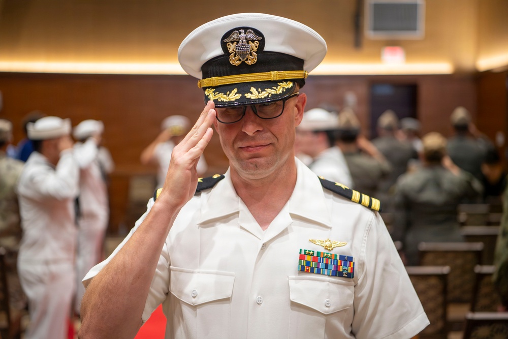 A U.S. Navy officer in white service dress uniform is rendering a salute during what appears to be a formal military ceremony. The officer wears a combination cover with gold oak leaf scrambled eggs indicating senior officer rank, and displays multiple service ribbons and aviator wings on his chest. In the background, other naval personnel in white uniforms are also visible, suggesting a change of command or similar formal event.