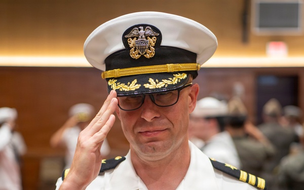A U.S. Navy officer in white service dress uniform is rendering a salute during what appears to be a formal military ceremony. The officer wears a combination cover with gold oak leaf scrambled eggs indicating senior officer rank, and displays multiple service ribbons and aviator wings on his chest. In the background, other naval personnel in white uniforms are also visible, suggesting a change of command or similar formal event.