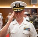 A U.S. Navy officer in white service dress uniform is rendering a salute during what appears to be a formal military ceremony. The officer wears a combination cover with gold oak leaf scrambled eggs indicating senior officer rank, and displays multiple service ribbons and aviator wings on his chest. In the background, other naval personnel in white uniforms are also visible, suggesting a change of command or similar formal event.