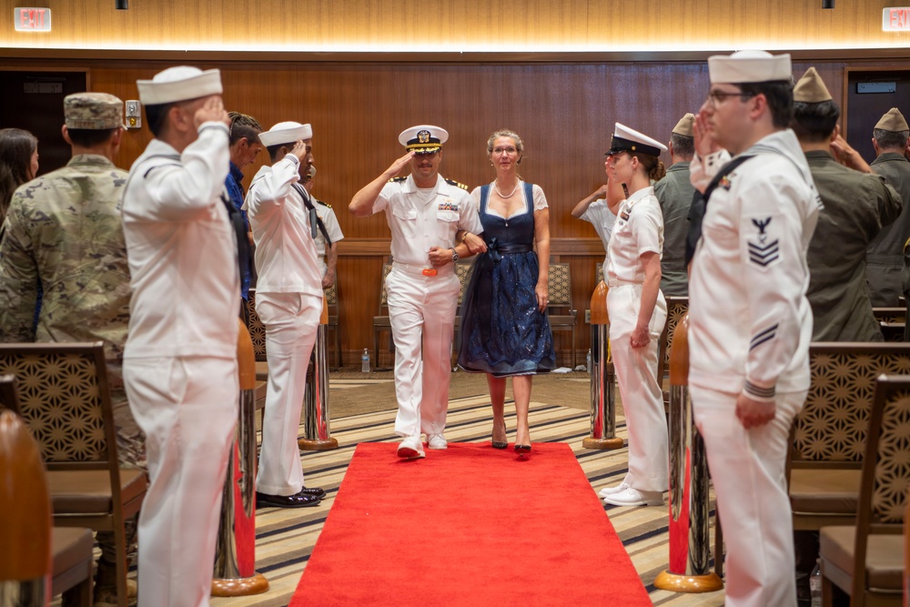 A U.S. Navy officer in dress white uniform walks down a red carpet alongside a woman in a blue dress during what appears to be a formal military ceremony, likely a change of command or retirement event. The officer is saluting while flanked by two rows of Navy enlisted personnel in dress whites who are also rendering salutes. Additional military personnel in various uniforms are visible in the background.