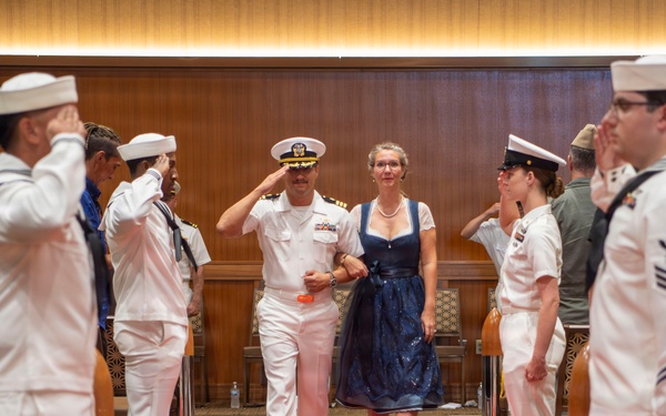 A U.S. Navy officer in dress white uniform walks down a red carpet alongside a woman in a blue dress during what appears to be a formal military ceremony, likely a change of command or retirement event. The officer is saluting while flanked by two rows of Navy enlisted personnel in dress whites who are also rendering salutes. Additional military personnel in various uniforms are visible in the background.