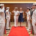 A U.S. Navy officer in dress white uniform walks down a red carpet alongside a woman in a blue dress during what appears to be a formal military ceremony, likely a change of command or retirement event. The officer is saluting while flanked by two rows of Navy enlisted personnel in dress whites who are also rendering salutes. Additional military personnel in various uniforms are visible in the background.