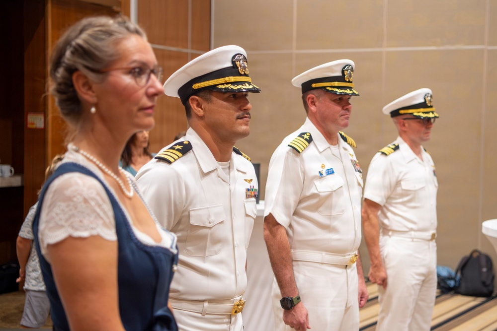 Three U.S. Navy officers in white service dress uniforms stand at attention in what appears to be an indoor ceremony or formal event. A civilian woman in a blue dress and white lace top stands in the foreground to the left. The officers display rank insignia and service ribbons consistent with senior Naval officer grades.
