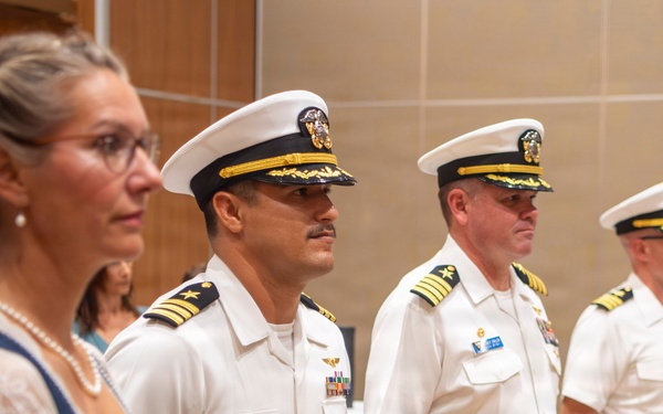 Three U.S. Navy officers in white service dress uniforms stand at attention in what appears to be an indoor ceremony or formal event. A civilian woman in a blue dress and white lace top stands in the foreground to the left. The officers display rank insignia and service ribbons consistent with senior Naval officer grades.