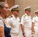 Three U.S. Navy officers in white service dress uniforms stand at attention in what appears to be an indoor ceremony or formal event. A civilian woman in a blue dress and white lace top stands in the foreground to the left. The officers display rank insignia and service ribbons consistent with senior Naval officer grades.
