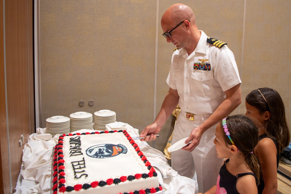 A U.S. Navy officer in white dress uniform is cutting a large decorated sheet cake at what appears to be a military unit celebration. Two young girls are watching closely as he prepares to serve slices. The cake is decorated with red and black frosting borders and features a squadron patch logo with the text 'STEEL DRAGONS'.