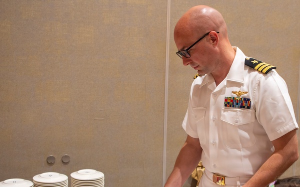 A U.S. Navy officer in white dress uniform is cutting a large decorated sheet cake at what appears to be a military unit celebration. Two young girls are watching closely as he prepares to serve slices. The cake is decorated with red and black frosting borders and features a squadron patch logo with the text 'STEEL DRAGONS'.