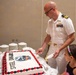 A U.S. Navy officer in white dress uniform is cutting a large decorated sheet cake at what appears to be a military unit celebration. Two young girls are watching closely as he prepares to serve slices. The cake is decorated with red and black frosting borders and features a squadron patch logo with the text 'STEEL DRAGONS'.