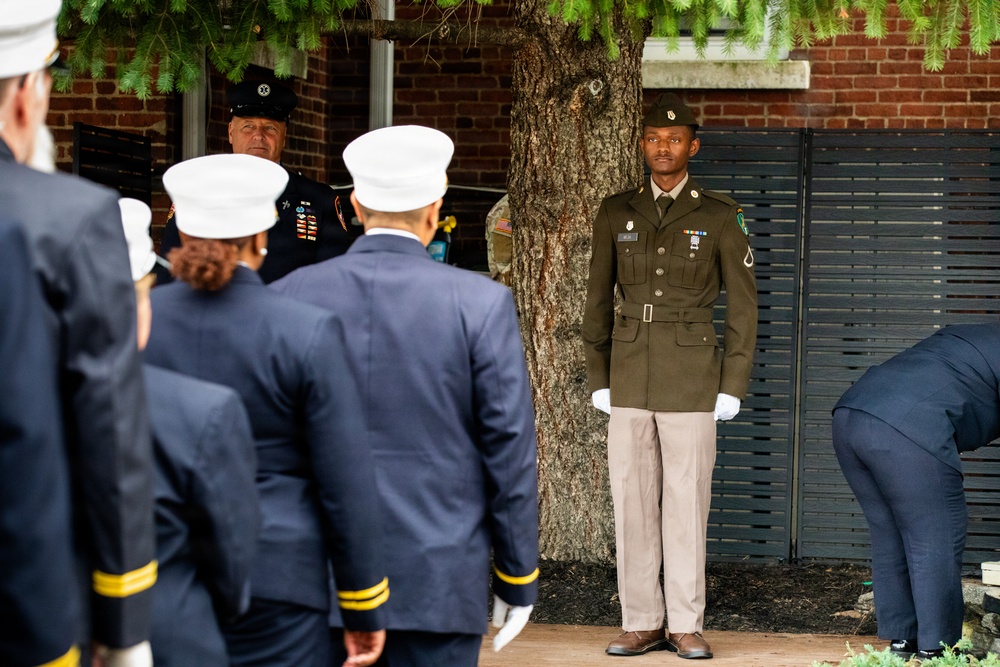 A formal military or ceremonial event is taking place outdoors near a brick building with a large tree. A young soldier in olive green Army dress uniform stands at attention wearing white gloves, while several individuals in navy blue uniforms with white caps — likely fire or police honor guard members — face away from the camera. A senior officer in a dark uniform with a black cap and medals is also visible in the background.