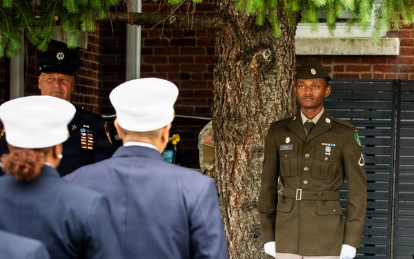 A formal military or ceremonial event is taking place outdoors near a brick building with a large tree. A young soldier in olive green Army dress uniform stands at attention wearing white gloves, while several individuals in navy blue uniforms with white caps — likely fire or police honor guard members — face away from the camera. A senior officer in a dark uniform with a black cap and medals is also visible in the background.