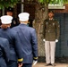 A formal military or ceremonial event is taking place outdoors near a brick building with a large tree. A young soldier in olive green Army dress uniform stands at attention wearing white gloves, while several individuals in navy blue uniforms with white caps — likely fire or police honor guard members — face away from the camera. A senior officer in a dark uniform with a black cap and medals is also visible in the background.