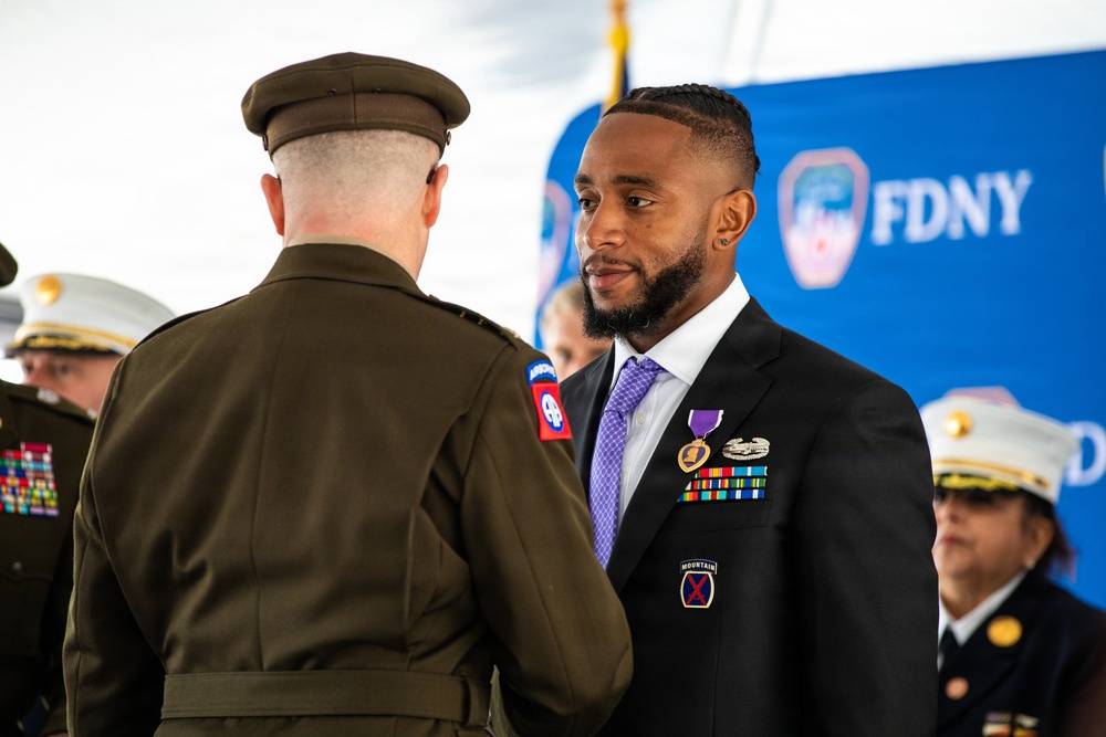 A formal military/civic ceremony is taking place in front of an FDNY backdrop. A civilian man in a black suit is wearing a Purple Heart medal and military service ribbons, suggesting he is a veteran being honored. A uniformed military officer with an 82nd Airborne Division patch is facing him, likely presenting or acknowledging the award.