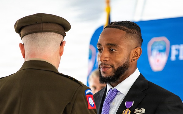 A formal military/civic ceremony is taking place in front of an FDNY backdrop. A civilian man in a black suit is wearing a Purple Heart medal and military service ribbons, suggesting he is a veteran being honored. A uniformed military officer with an 82nd Airborne Division patch is facing him, likely presenting or acknowledging the award.