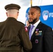 A formal military/civic ceremony is taking place in front of an FDNY backdrop. A civilian man in a black suit is wearing a Purple Heart medal and military service ribbons, suggesting he is a veteran being honored. A uniformed military officer with an 82nd Airborne Division patch is facing him, likely presenting or acknowledging the award.