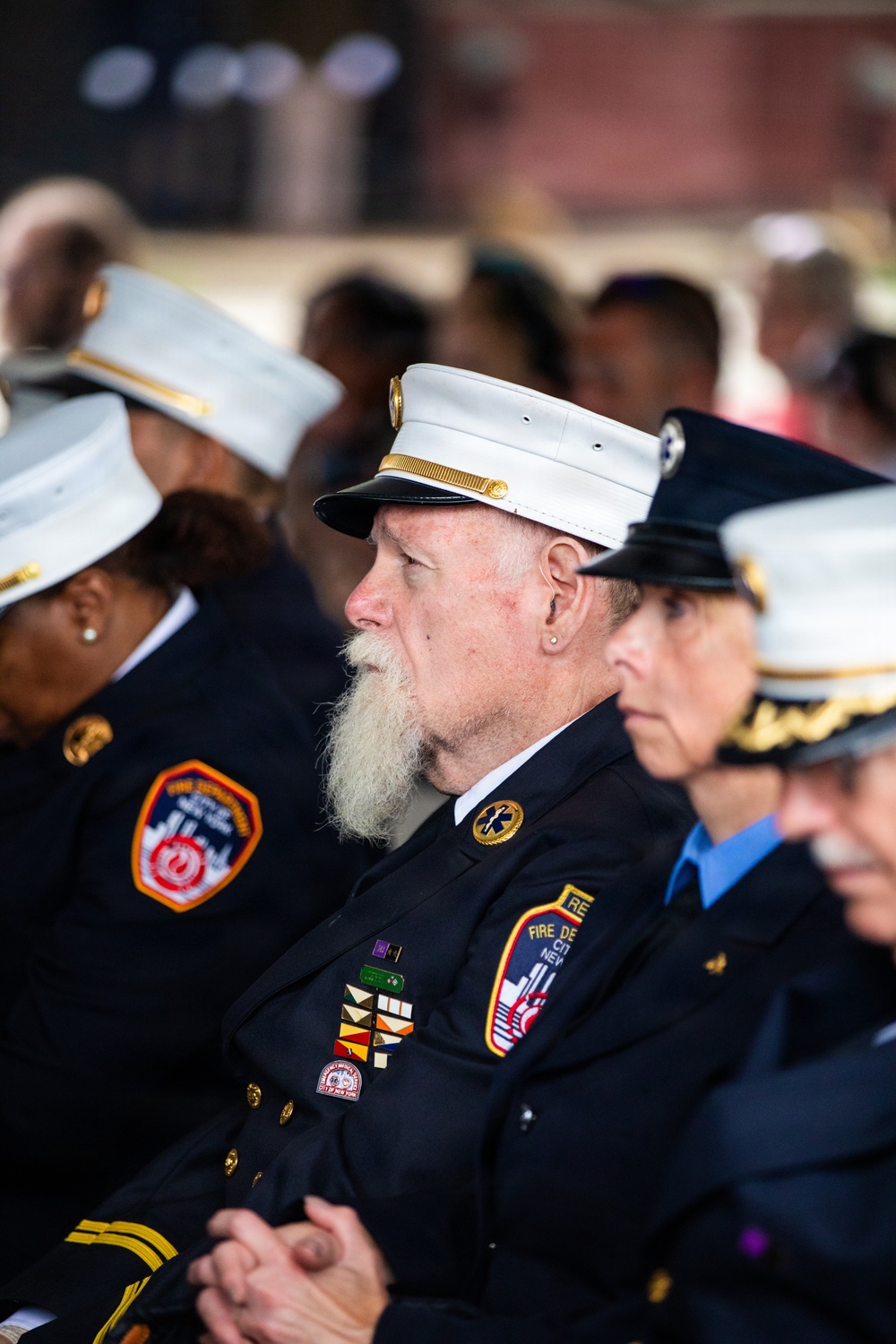 The image shows several uniformed members of the FDNY (Fire Department City of New York) seated at what appears to be a formal ceremony or memorial event. The central figure is an older man with a white beard wearing a white-topped dress uniform cap and a dark navy uniform adorned with service ribbons, medals, and an EMS star-of-life pin. Multiple FDNY patches and insignia are clearly visible on the uniforms.